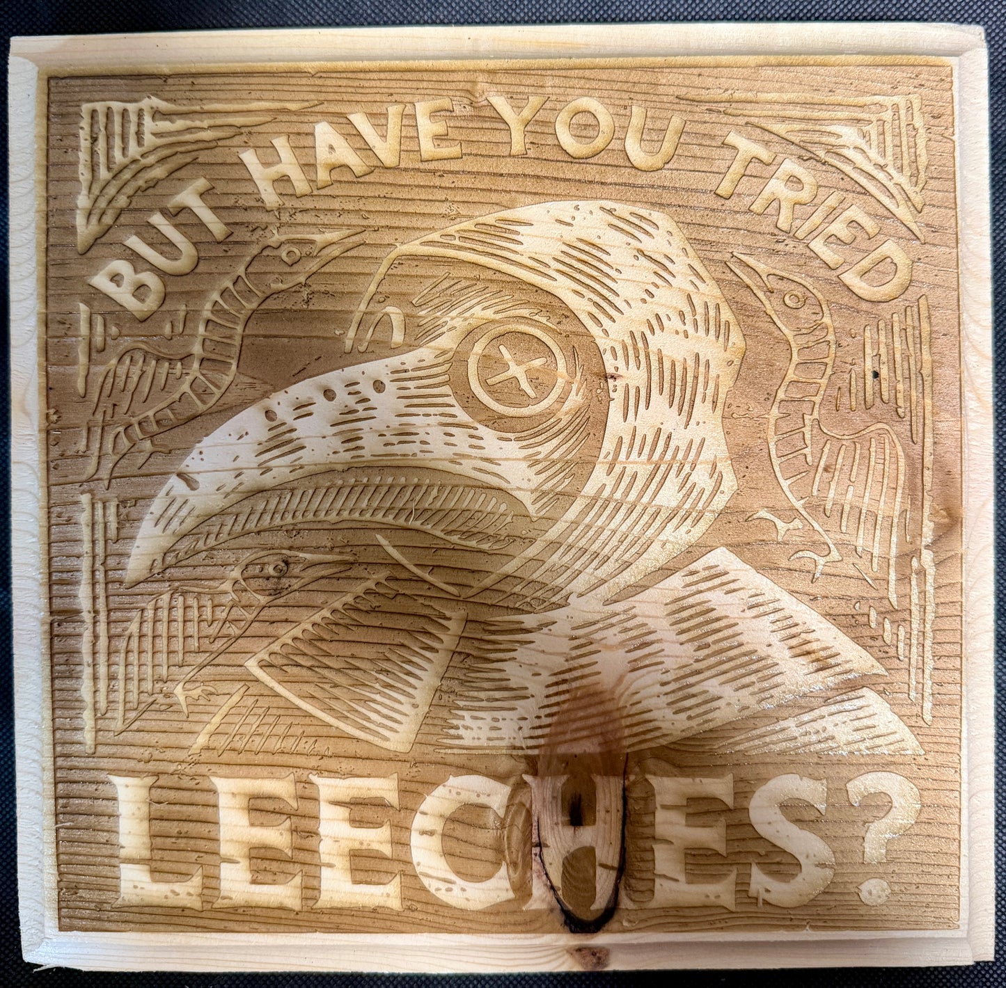 Wooden block with engraved plague doctor and text 'But have you tried leeches?'