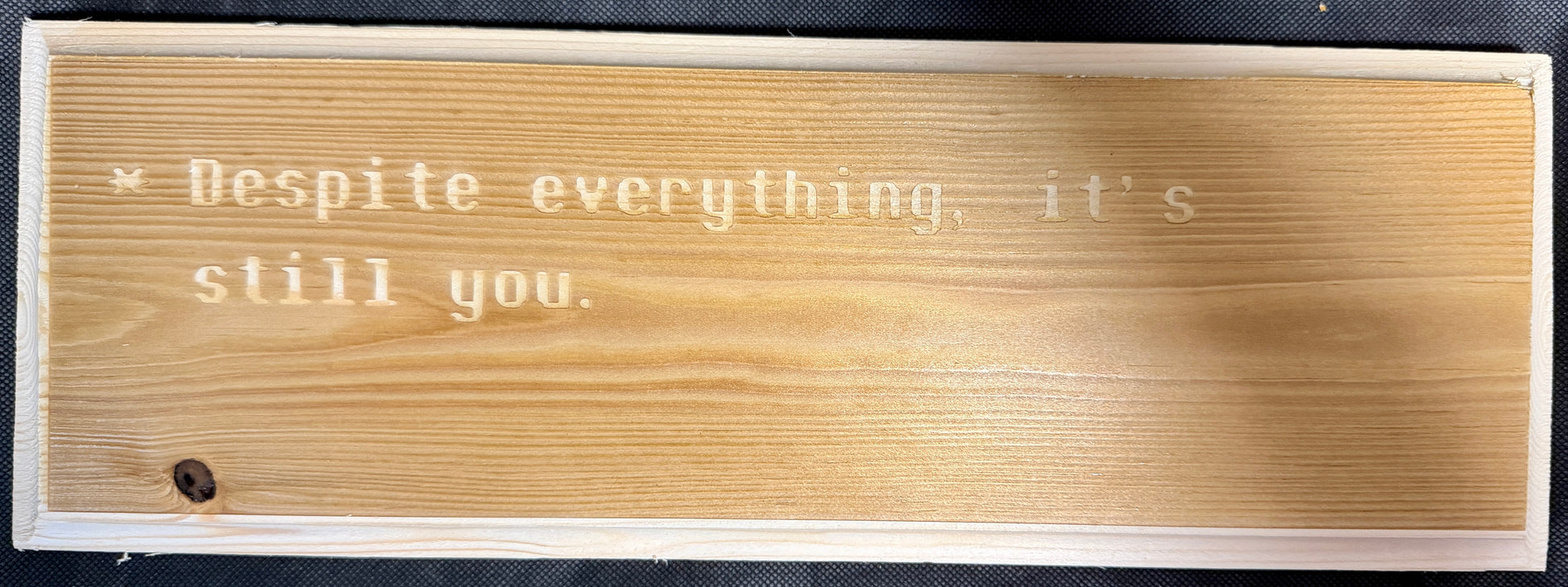 Wooden plaque with engraved text 'Despite everything, it's still you.' on a black background