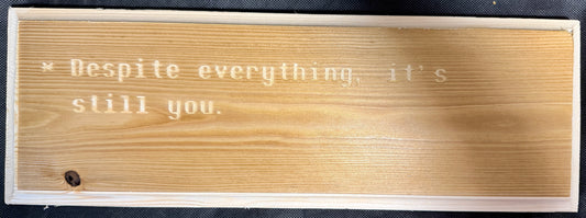 Wooden plaque with engraved text 'Despite everything, it's still you.' on a black background