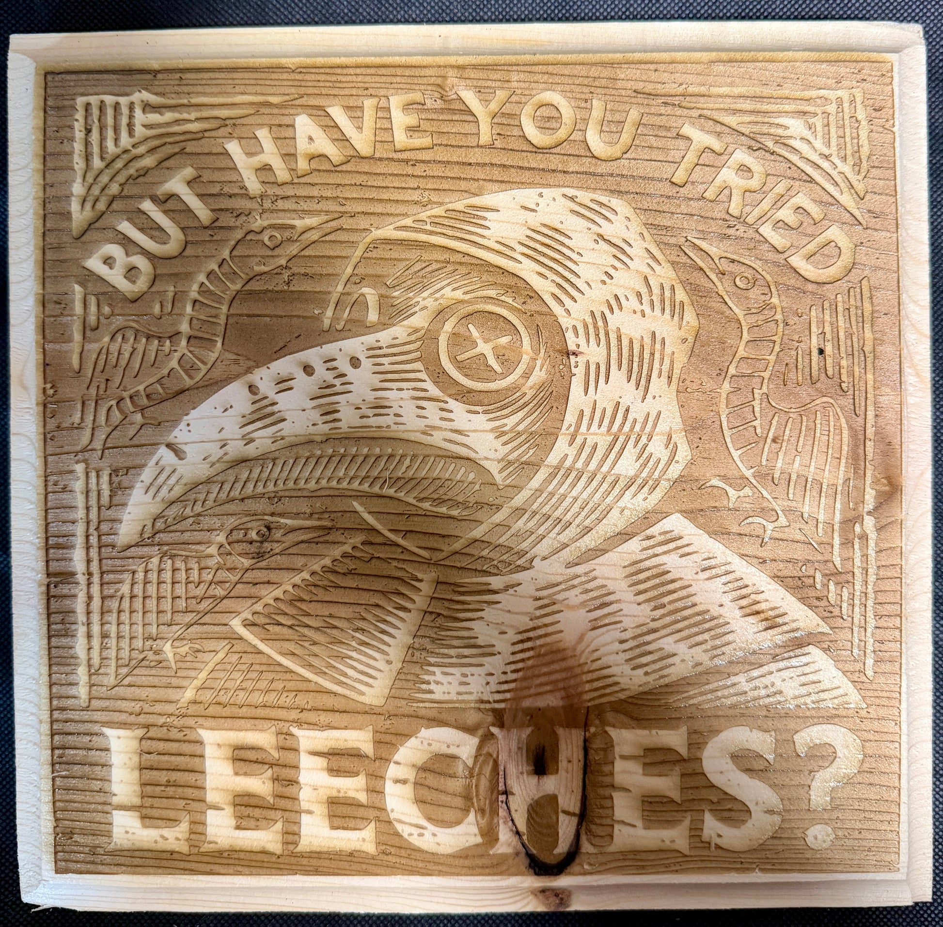 Wooden block with engraved plague doctor and text 'But have you tried leeches?'