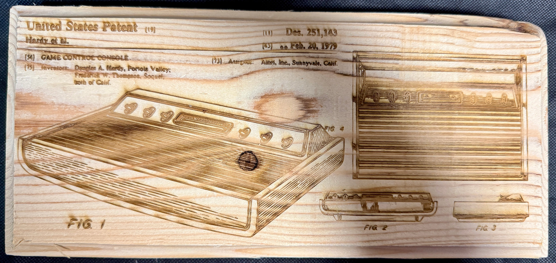 Wooden plaque with engraved design of a vintage video game console, resembling a patent illustration.